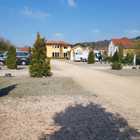 Ein ruhiger Parkplatz mit Kies und einigen Bäumen. Im Hintergrund sind mehrere Gebäude und Fahrzeuge zu sehen.