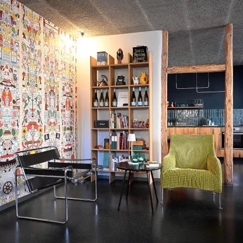Ein modernes Wohnzimmer mit buntem Wanddesign und einem Bücherregal. Im Raum stehen ein schwarzer Stuhl und ein grüner Sessel.