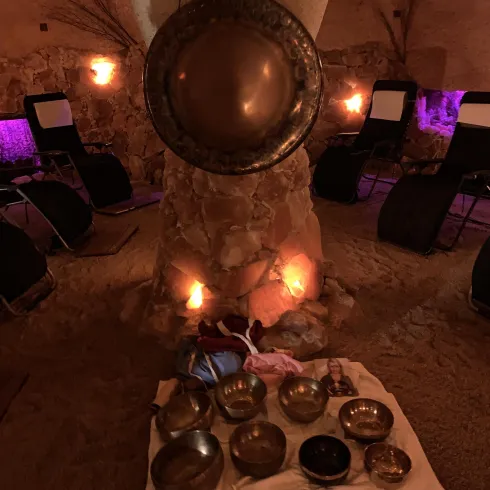 Ein entspannender Raum mit einer Sandfläche und leuchtenden Wänden. In der Mitte stehen verschiedene Klangschalen auf einem Tuch.