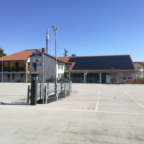 Ein Parkplatz mit einer großen, leeren Fläche. Im Hintergrund sind mehrere Gebäude mit Sonnenkollektoren zu sehen.