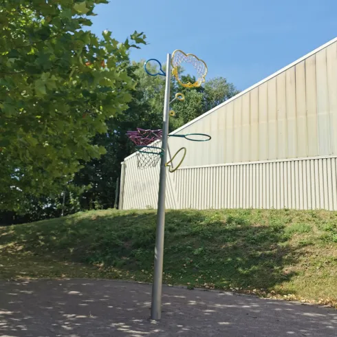 Eine Sportstätte mit einem Basketballkorb vor einer Wand. Der Himmel ist klar und es gibt Bäume in der Nähe.