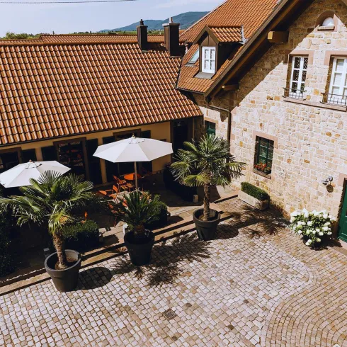 Ein malerischer Innenhof mit gepflastertem Boden und Pflanzen in Töpfen. Zwei Sonnenschirme bieten Schatten für die Sitzgelegenheiten.