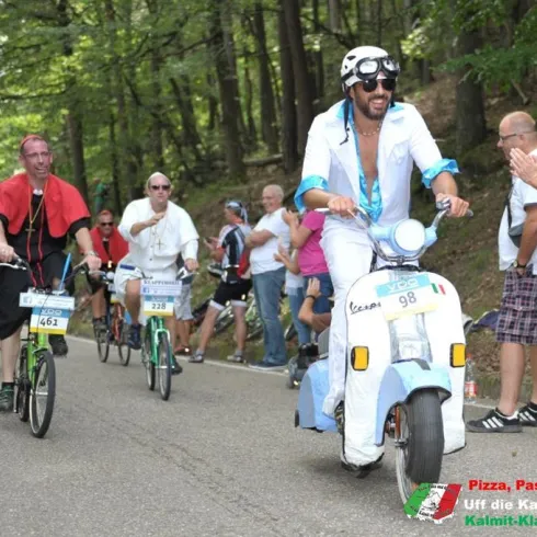 Eine bunte Veranstaltung mit Radfahrern in sportlicher Kleidung, die eine Straße entlangfahren. Im Vordergrund fährt jemand auf einem Moped in auffälligem Outfit.