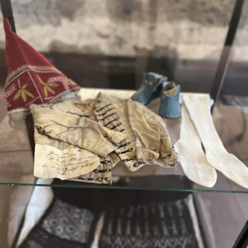 Auf dem Bild sind historische Textilien und Accessoires in einem Glasdisplay zu sehen. Dazu gehören Handschuhe, Socken und eine Kopfbedeckung mit bunten Mustern.