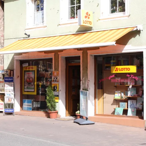 Ein kleiner Laden mit einem gelben Sonnenschutz und großen Schaufenstern. Vor dem Geschäft stehen Pflanzen und Werbetafeln.