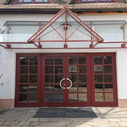 Ein Gebäude mit einer weißen Fassade und großen roten Fenstern. Über dem Eingang befindet sich eine markante Vordachkonstruktion aus Glas und Metall.