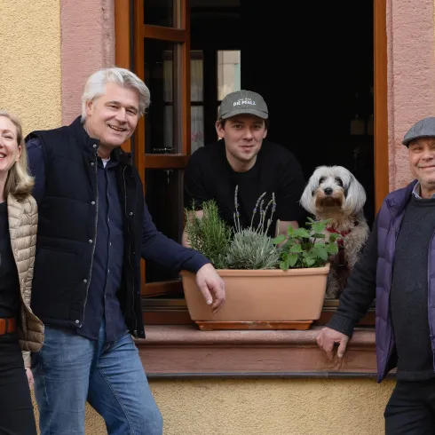 Eine Gruppe von vier Personen steht vor einem Fenster mit Pflanzkästen. Ein Hund schaut neugierig aus dem Fenster.
