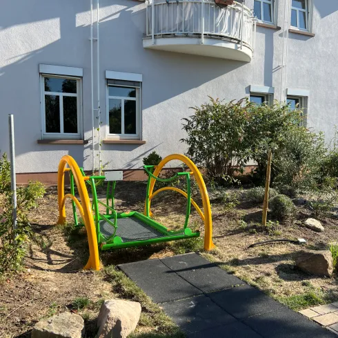 Ein farbenfrohes Spielgerät steht im Garten vor einem Wohnhaus. Der Spielplatz besteht aus einer grünen Fläche mit gelben Akzenten und ist von Sträuchern umgeben.