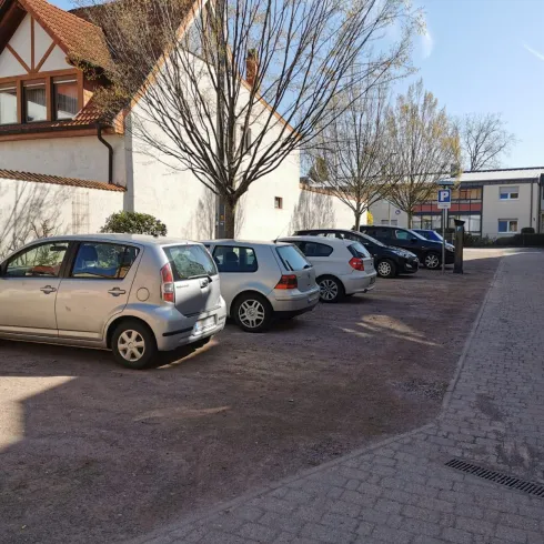 Ein ruhiger Parkplatz mit mehreren Autos und einem gepflasterten Weg. Im Hintergrund sind Bäume und Gebäude sichtbar.