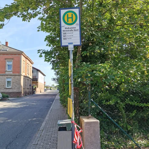 Eine Bus-Haltestelle steht an einer ruhigen Straße, umgeben von Bäumen. Das Schild zeigt die Abfahrtszeiten und Liniennummern an.