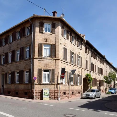Ein historisches Gebäude an einer Straßenecke mit charakteristischen Fensterläden. Die Fassade ist aus Sandstein, umgeben von einer ruhigen Straße.