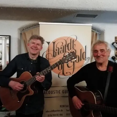 Zwei Musiker stehen nebeneinander und halten akustische Gitarren. Im Hintergrund ist ein Banner mit dem Schriftzug "Lizard of Gold" zu sehen.