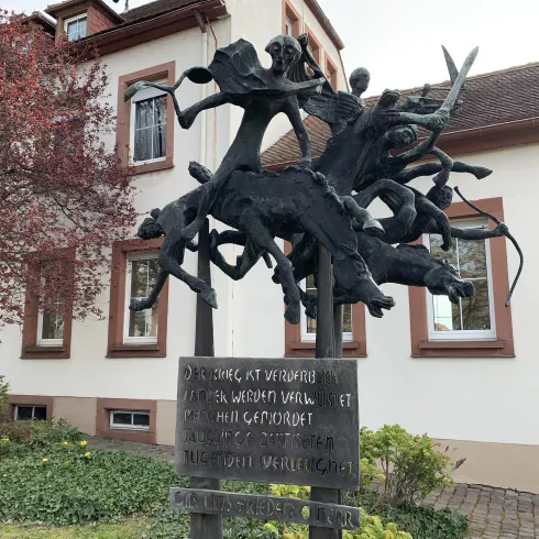 Eine kreative Skulptur mit dynamischen Figuren aus Bronze, die miteinander interagieren. Im Hintergrund steht ein Gebäude mit klassischen Fenstern und Bäumen.