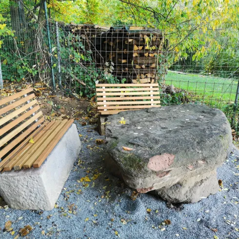 Zwei Bänke, eine aus Holz und eine aus Stein, stehen auf einem Kiesplatz umgeben von Bäumen. Im Hintergrund sind Holzstämme und ein grünes Feld zu sehen.