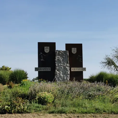 Ein monumentales Schild mit den Namen "Hainamt" und "Kirchweiler". Es ist von üppigem Grün und Blumen umgeben, unter einem klaren blauen Himmel.