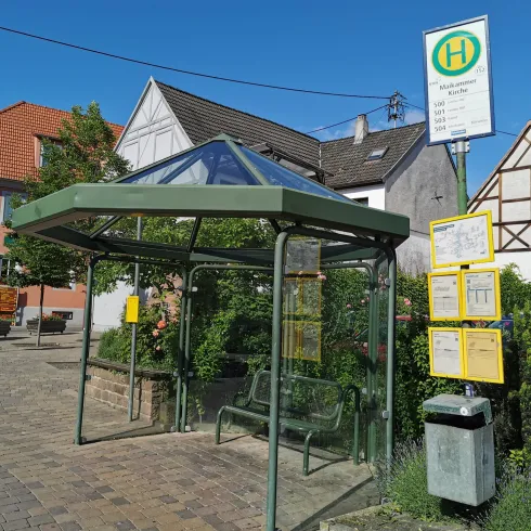 Eine moderne Bushaltestelle mit Glaskonstruktion und einer Informationsstelle. Im Hintergrund sind traditionelle Häuser und ein blauer Himmel zu sehen.