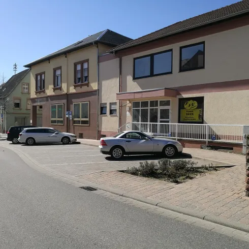 Eine ruhige Straße mit Häusern auf der linken Seite. Geparkte Autos stehen auf einem Platz vor den Gebäuden.