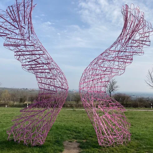 Eine kunstvolle Skulptur in Form von Flügeln steht in einer grünen Wiese. Der Himmel ist teilweise bewölkt und zeigt einen Hauch von Blau.