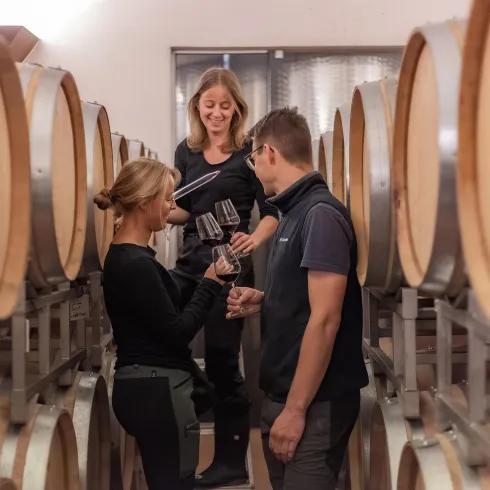 Drei Personen stehen in einem Weinkeller zwischen Eichenfässern. Sie halten Gläser und scheinen Wein zu probieren und zu diskutieren.