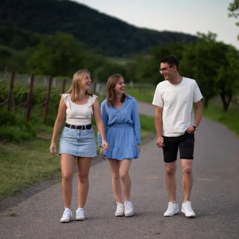 Drei junge Menschen spazieren lächelnd auf einem Weg. Die Umgebung ist grün und hügelig, ideal für einen entspannten Tag im Freien.