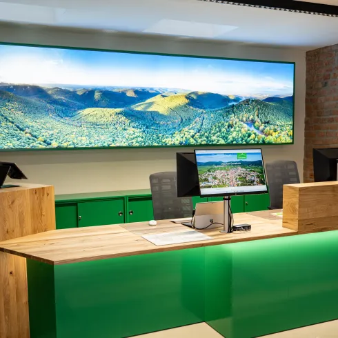 Ein modernes Empfangsbereich mit einem großen Bildschirm, der eine Landschaft zeigt. Der Tisch ist aus Holz und grünen Elementen gestaltet.