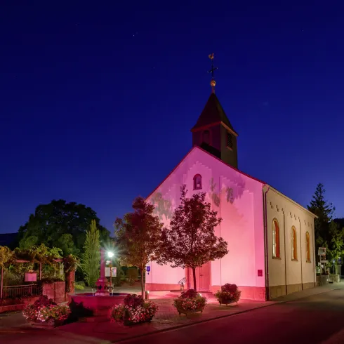 Eine schön beleuchtete Kirche steht in der Dämmerung. Bäume und Pflanzen umgeben das Gebäude in einer friedlichen Umgebung.