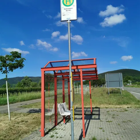 Eine Bushaltestelle mit rotem Überstand und zwei Sitzplätzen. Der Himmel ist blau mit einigen Wolken.