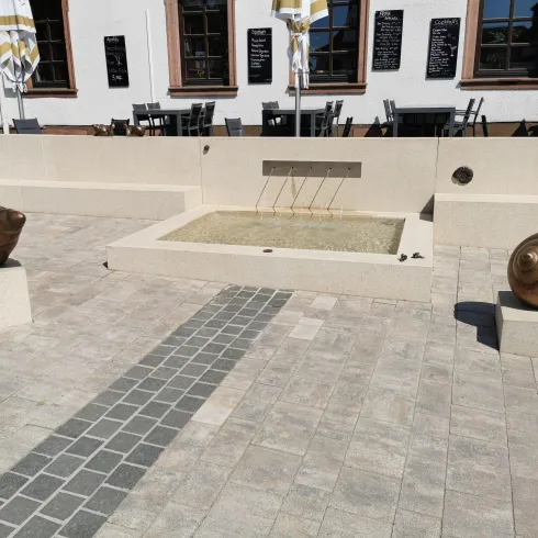 Eine von zwei Schneckenformen umgebene Brunnenanlage in einem öffentlichen Platz. Im Hintergrund sind Tische und Stühle eines Cafés zu sehen.