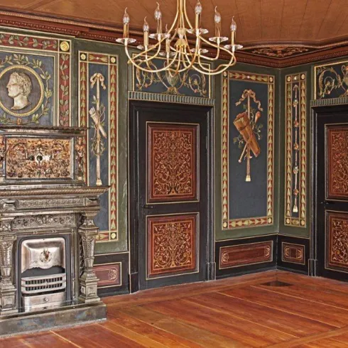 Ein historischer Raum mit kunstvoll verzierten Wänden und einem eleganten alten Kamin. Eine beeindruckende Deckenleuchte ergänzt die antike Atmosphäre.