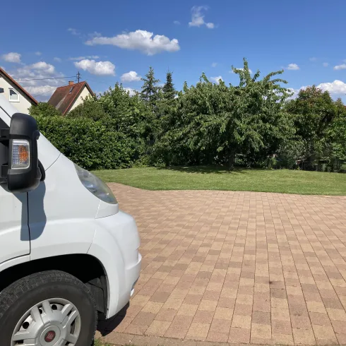 Ein weißes Fahrzeug steht auf einem gepflasterten Parkplatz. Im Hintergrund sind Bäume und ein klarer blauer Himmel zu sehen.