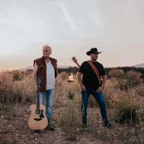 Ein Mann mit grauen Haaren steht mit einer Gitarre im Hintergrund. Er hat einen nachdenklichen Ausdruck und trägt ein einfaches schwarzes Oberteil.