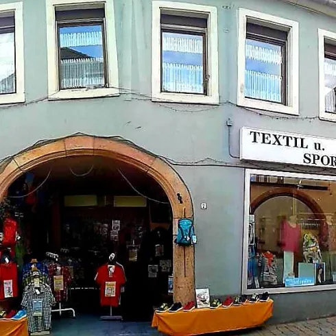 Ein Geschäft mit einem großen Schaufenster, das Sport- und Textilartikel verkauft. Die Fassade des Gebäudes ist freundlich und einladend gestaltet.