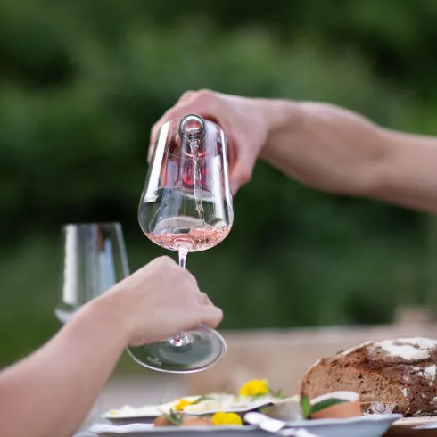 Eine Person gießt Roséwein in ein Glas. Im Hintergrund sieht man eine rustikale Brotlaib und Blumen auf dem Tisch.