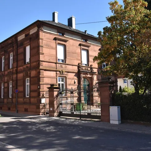 Ein historisches gemauertes Gebäude mit grünem Eingang und Tor. Vor dem Haus stehen Bäume und Blumen.