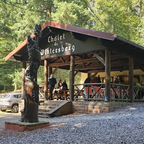 Ein Holzchalet im Wald mit der Aufschrift "Chalet du Wintersberg". Einige Besucher sind draußen versammelt, umgeben von grünen Bäumen.