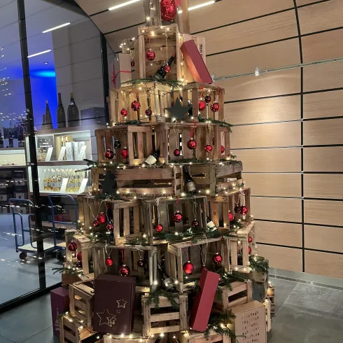 Ein festlich geschmückter Weihnachtsbaum aus Holzkisten, geschmückt mit roten Kugeln und Lichtern. Der Baum steht in einem modernen Raum mit großen Fenstern.