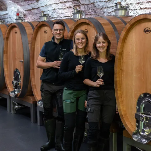Drei Personen stehen lächelnd vor großen Holzfässern in einem Weinkeller. Alle halten ein Glas in der Hand und tragen schwarze Kleidung.