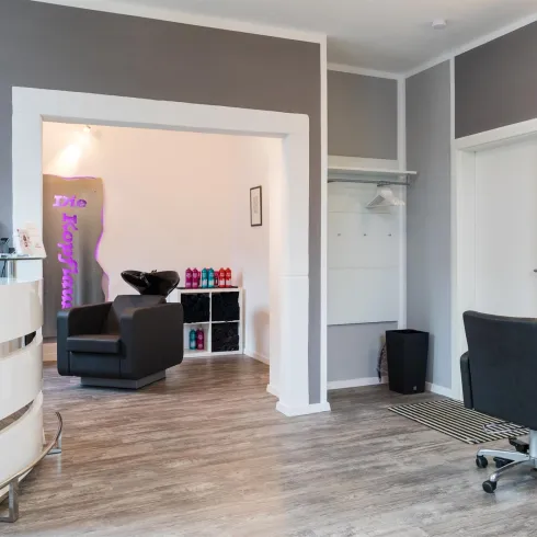 Ein eleganter Friseursalon mit modernen Stühlen und einem großen Spiegel. Die Wände sind hellgrau, und es gibt eine gemütliche Sitzecke.