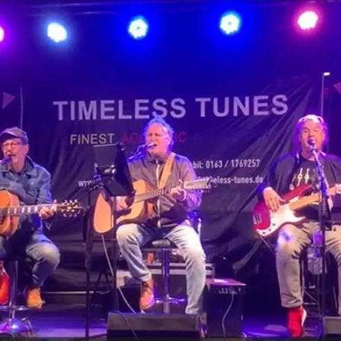 Ein Mann mit grauen Haaren steht mit einer Gitarre im Hintergrund. Er hat einen nachdenklichen Ausdruck und trägt ein einfaches schwarzes Oberteil.