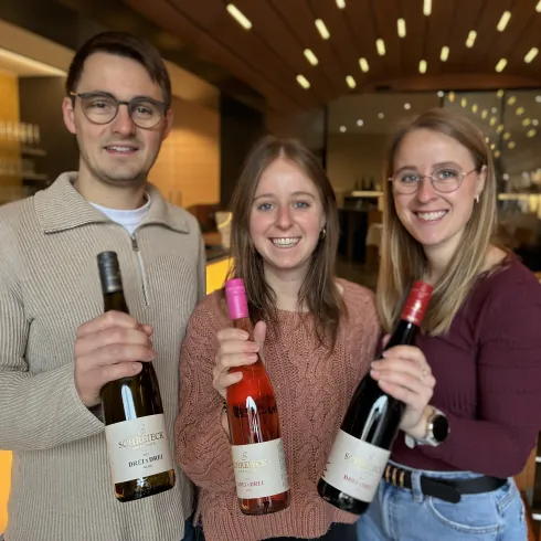 Drei Personen stehen in einem Weinlokal und halten Weinflaschen in der Hand. Sie lächeln und scheinen sich zu freuen.