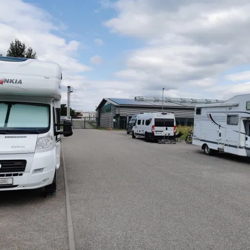Ein Parkplatz mit mehreren Wohnmobilen unter einem bewölkten Himmel. Im Hintergrund ist ein Gebäude sichtbar.