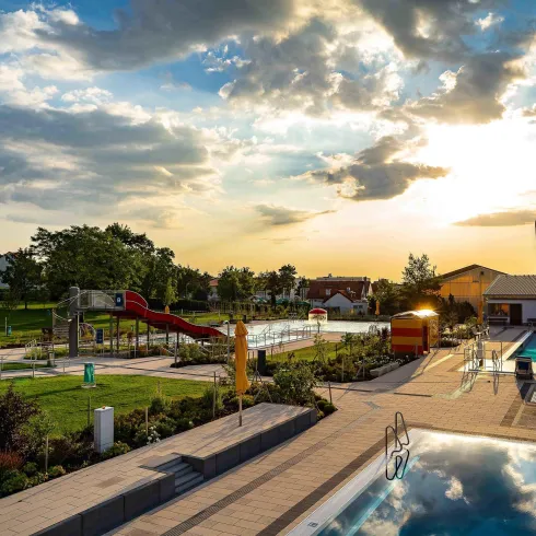 Ein schöner Außenpool mit rutschigen Anlagen und einer weitläufigen Gartenlandschaft. Die Sonne geht unter und der Himmel ist teilweise bewölkt.