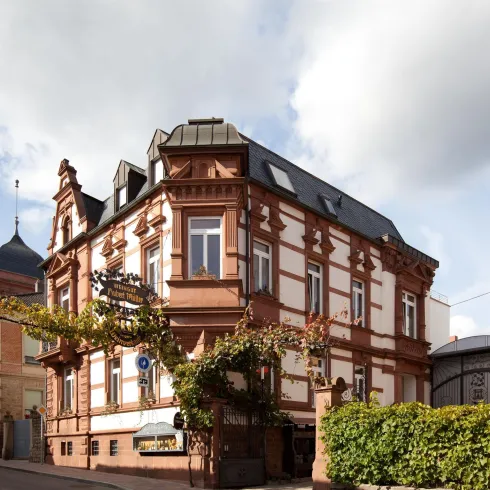 Ein historisches Gebäude mit rotem Sandstein und vielen Fenstern. Im Vordergrund sind grüne Pflanzen und die Straße sichtbar.