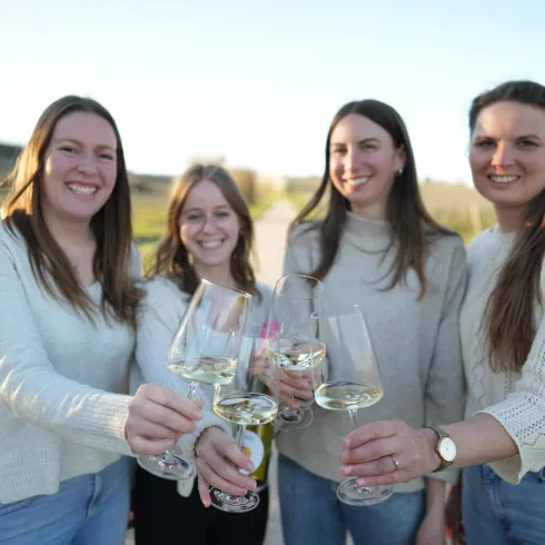 Vier Frauen stehen im Freien und halten Gläser mit Getränken. Sie lächeln und scheinen einen fröhlichen Moment zu genießen.