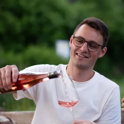Ein Mann gießt Roséwein in ein Glas. Im Hintergrund sind grüne Bäume zu sehen.