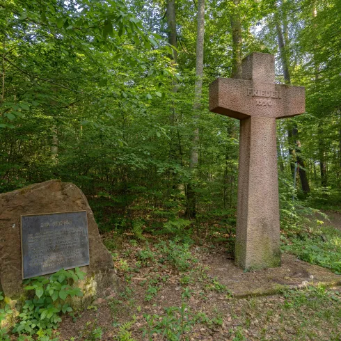 Ein Gedenkkreuz aus Stein steht in einem grünen Wald. Neben dem Kreuz befindet sich ein Gedenkstein mit einer Tafel.