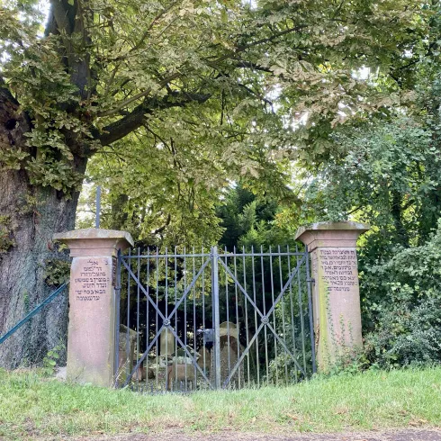 Ein schmaler Eingang mit einem schwarzen Gitterzaun, umgeben von Bäumen und Grün. Auf den Säulen stehen Inschriften, die dem Ort Bedeutung verleihen.