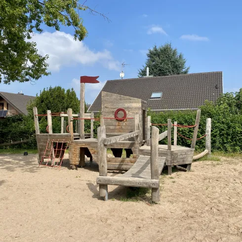 Ein Spielplatz aus Holz mit verschiedenen Strukturen zum Klettern und Spielen. Im Hintergrund sind Bäume und ein Haus sichtbar.
