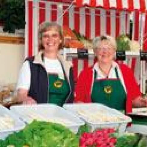 Zwei Frauen stehen an einem Marktstand mit frischem Gemüse. Der Stand ist mit roten und weißen Streifen dekoriert.