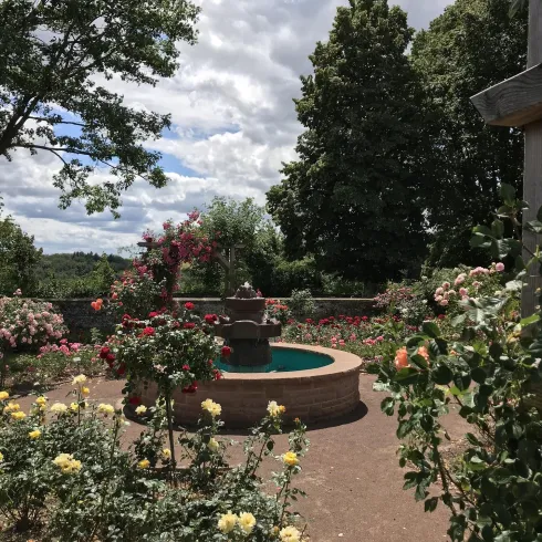 Ein schöner Garten mit vielen bunten Rosen und einem Brunnen in der Mitte. Der Himmel ist teils bewölkt und bietet einen malerischen Hintergrund.